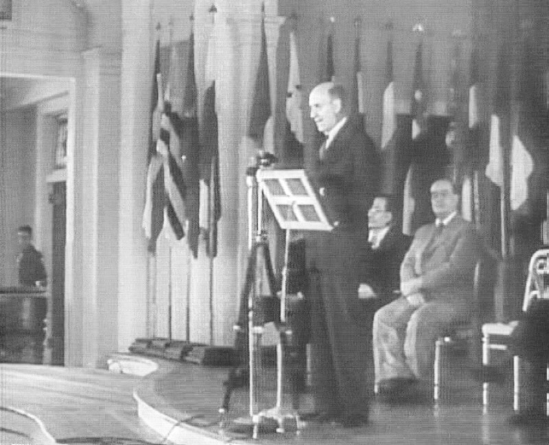 A film still of a man speaking at a lectern with a microphone, national flags behind him and two men seated beside him, at the opening of the Bretton Woods conference in July 1944.