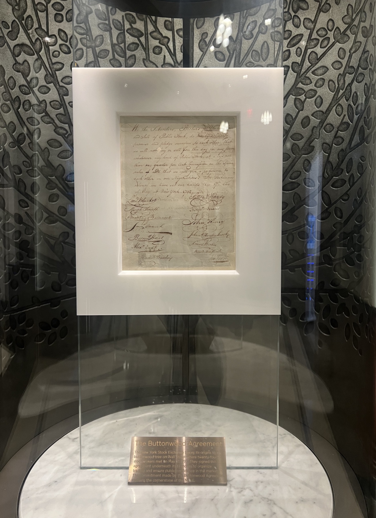The Buttonwood Agreement, a single handwritten sheet in a white mount inside a glass case on a marble plinth at the New York Stock Exchange, with a brass plaque below it.