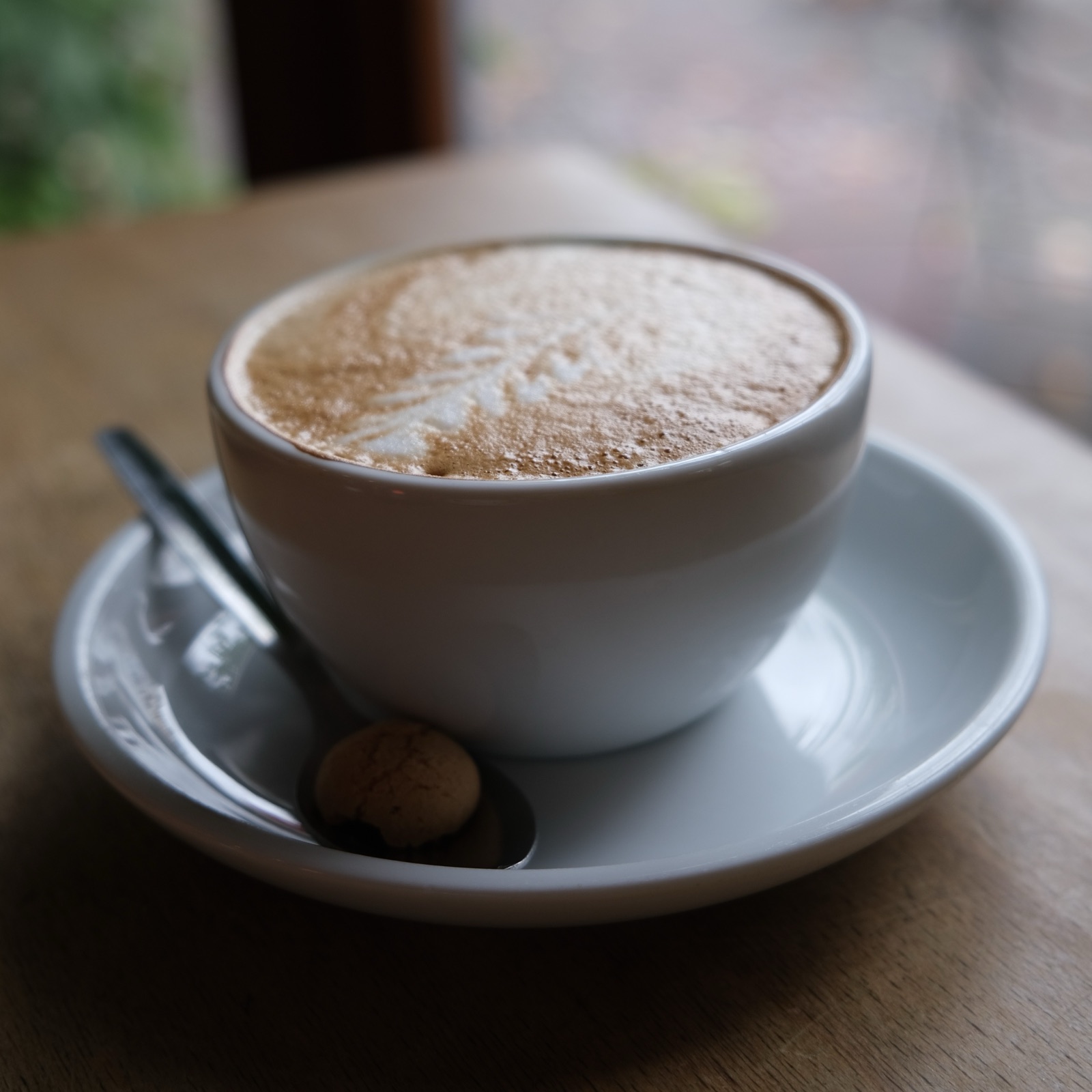 A cappuccino in a white cup on a saucer, with a spoon and a small biscuit, on a wooden table by a window.