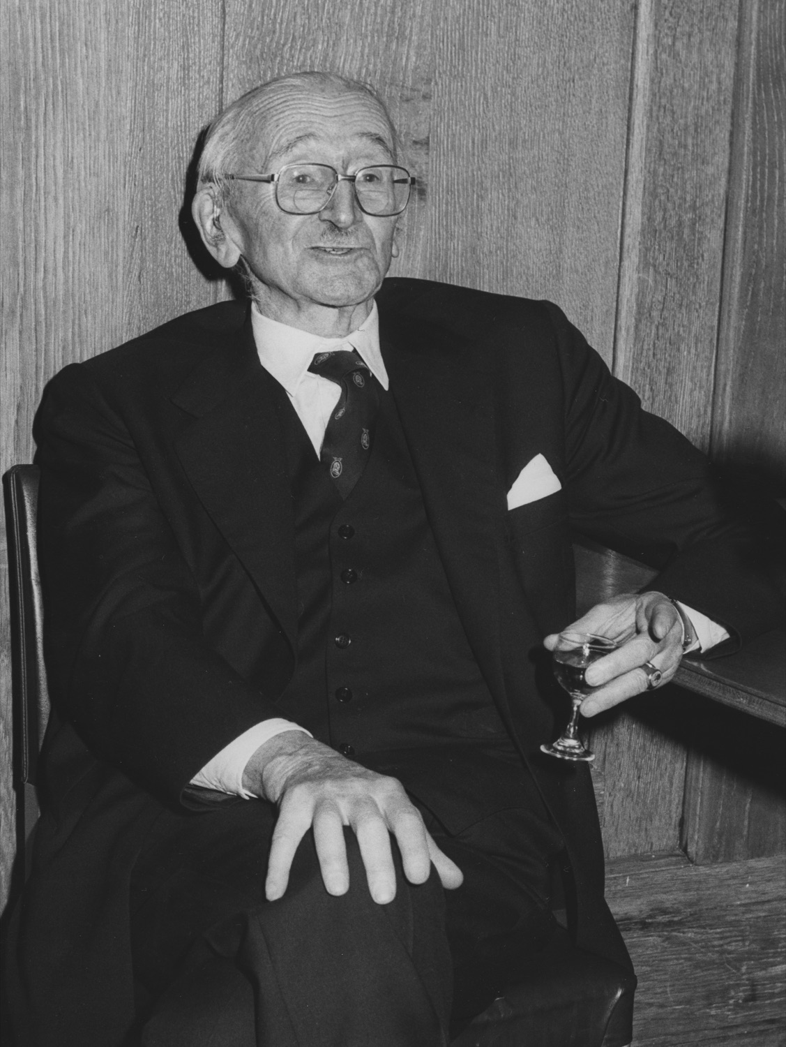 Friedrich Hayek in old age, seated in a wood-panelled room in a three-piece suit and glasses, holding a small glass, at the London School of Economics in 1981.