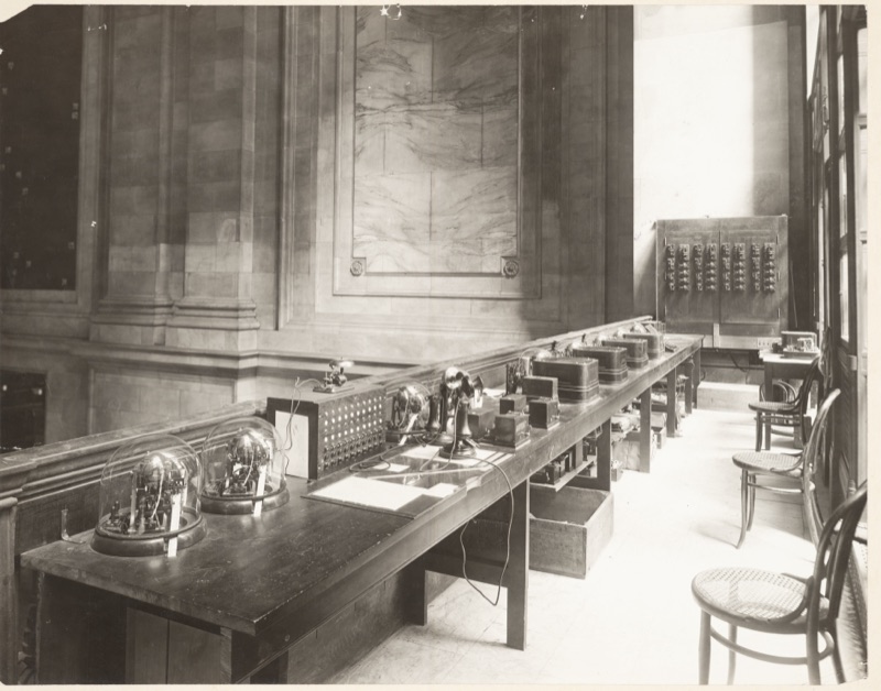 A long dark wooden desk in a high stone hall holding ticker machines under glass domes, telephones and a switchboard, with a wall panel of switches behind and empty chairs beside it.