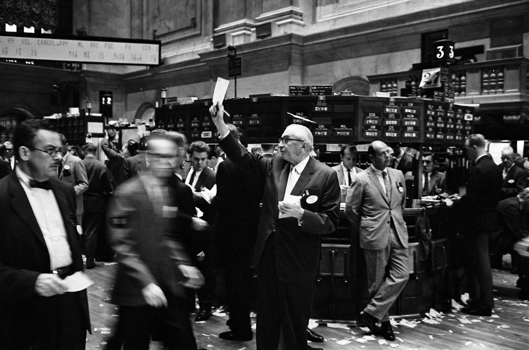 The floor of the New York Stock Exchange in 1963. In the centre an older man in a suit and glasses holds a paper slip high above his head with one arm, looking to his right. Brokers crowd around him and trading posts with numbered signs stand behind.