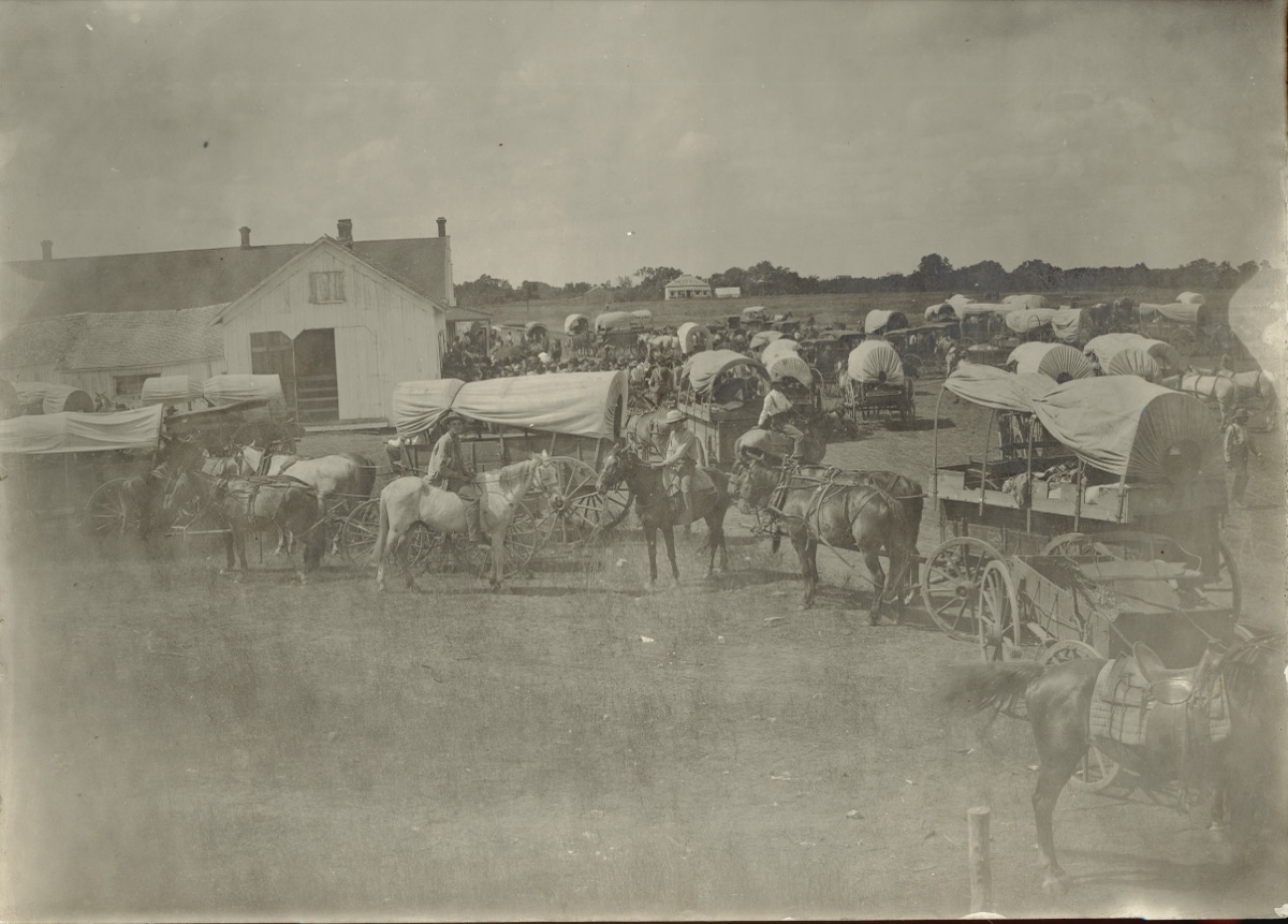 Covered wagons, teams of horses and a crowd gathered around a building near Fort Sill in July 1901.