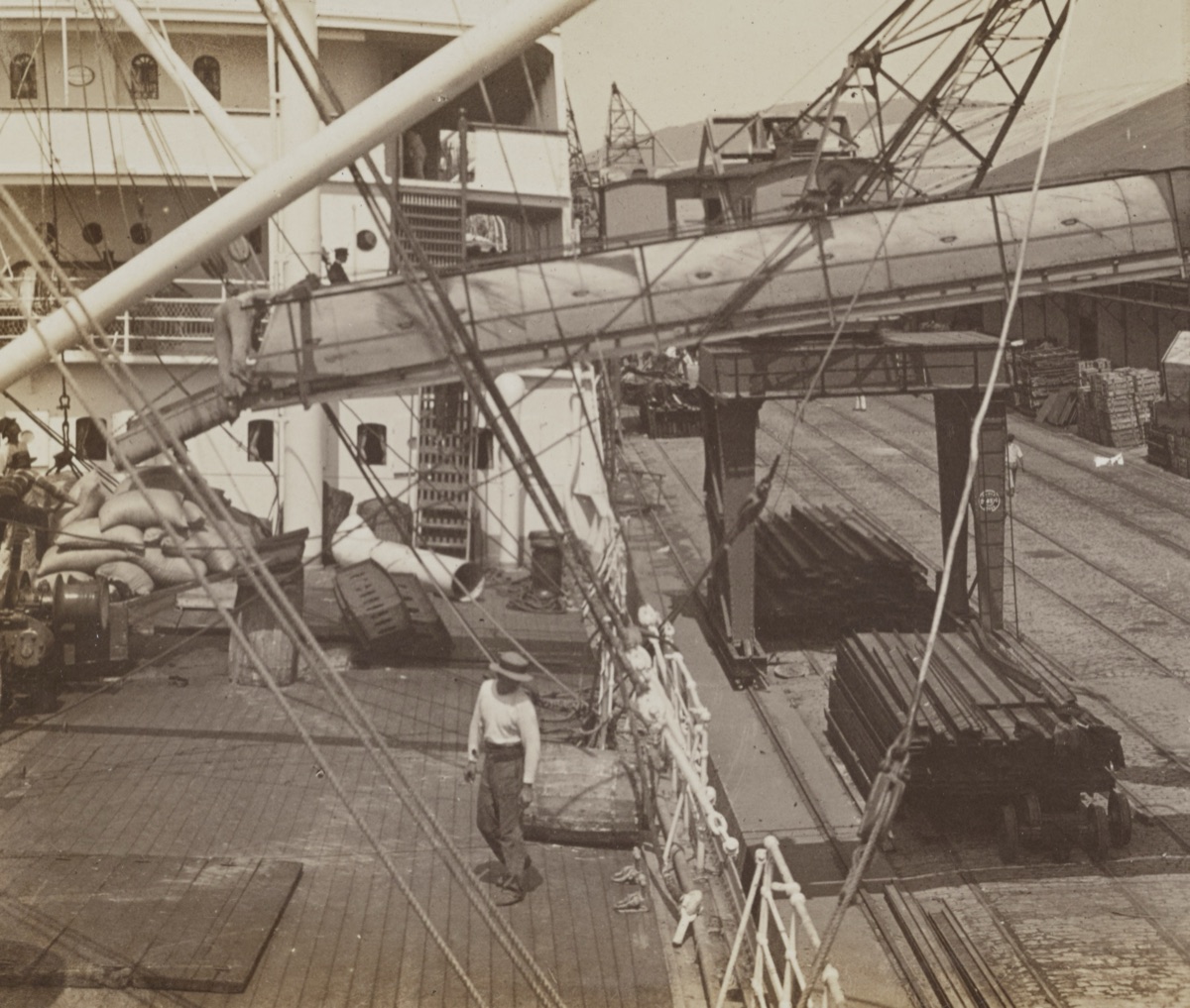 Sepia stereograph, one half: a conveyor chute runs from the quay onto a steamship, bags of coffee piled on its deck, a man in a hat standing by, rail tracks and timber on the quay.