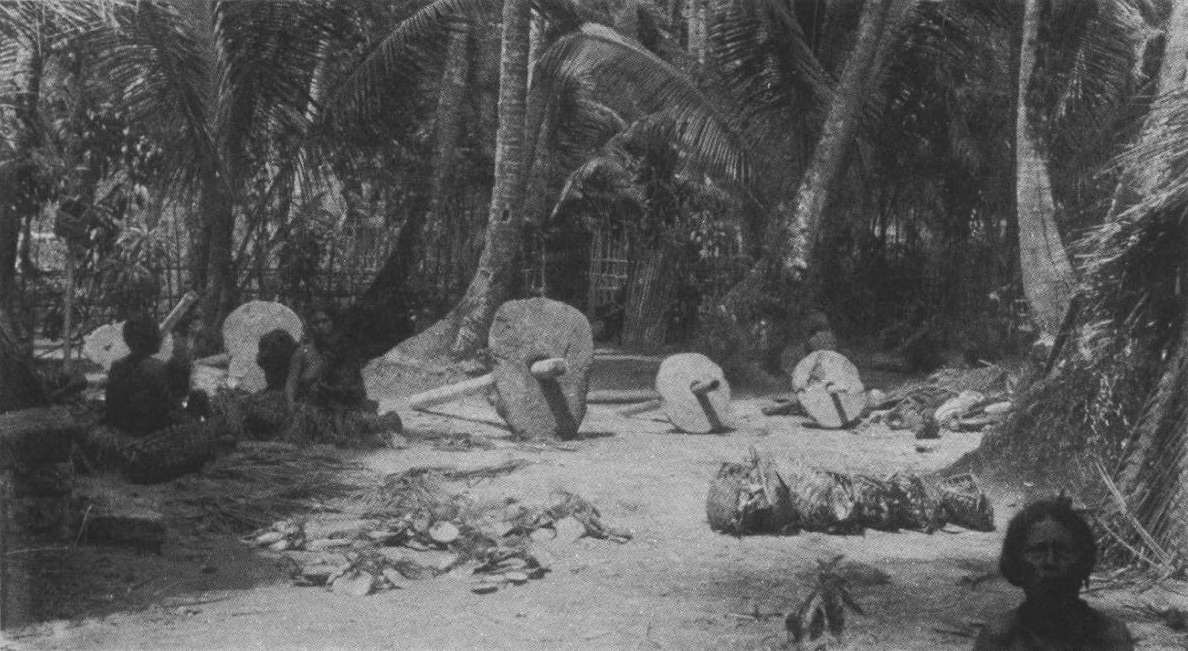 Under coconut palms on Uap, several large round stone discs stand on edge with wooden poles through their centres. People sit in the shade beside them.